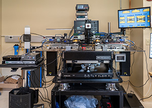 Load Pull System. Complex semiconductor test equipment in a lab. A large machine with metallic components, wires, and multiple screens is central.